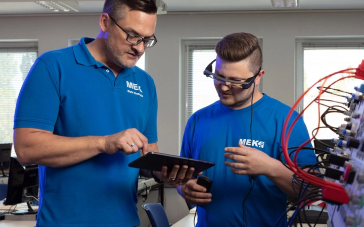 Ausbilder und Teilnehmer schauen auf ein Tablet, während der Teilnehmer eine spezielle VR-Brille trägt, in die Arbeitsanweisungen und technische Informationen eingeblendet werden.