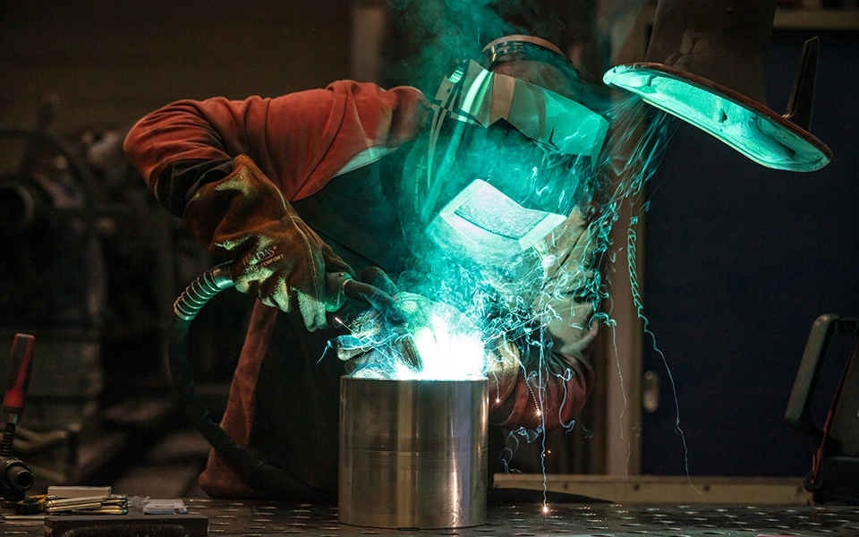 Person schweißt unter Verwendung von Schutzkleidung, während die Flamme des Schweißgeräts gut sichtbar ist.