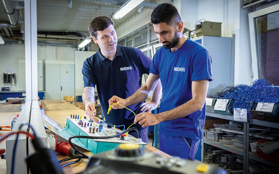 Ein Ausbilder zeigt einem Teilnehmer einer Elektro-Ausbildung in der Werkstatt, wie man Elektroinstallationen durchführt.