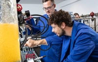 Auszubildender beugt sich nach vorne und zeigt auf eine Komponente am Hydraulikversuchsstand, während er die Funktionsweise hydraulischer Systeme untersucht.