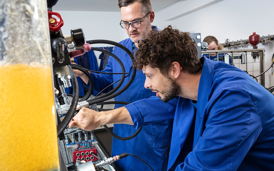 Auszubildender beugt sich nach vorne und zeigt auf eine Komponente am Hydraulikversuchsstand, während er die Funktionsweise hydraulischer Systeme untersucht.