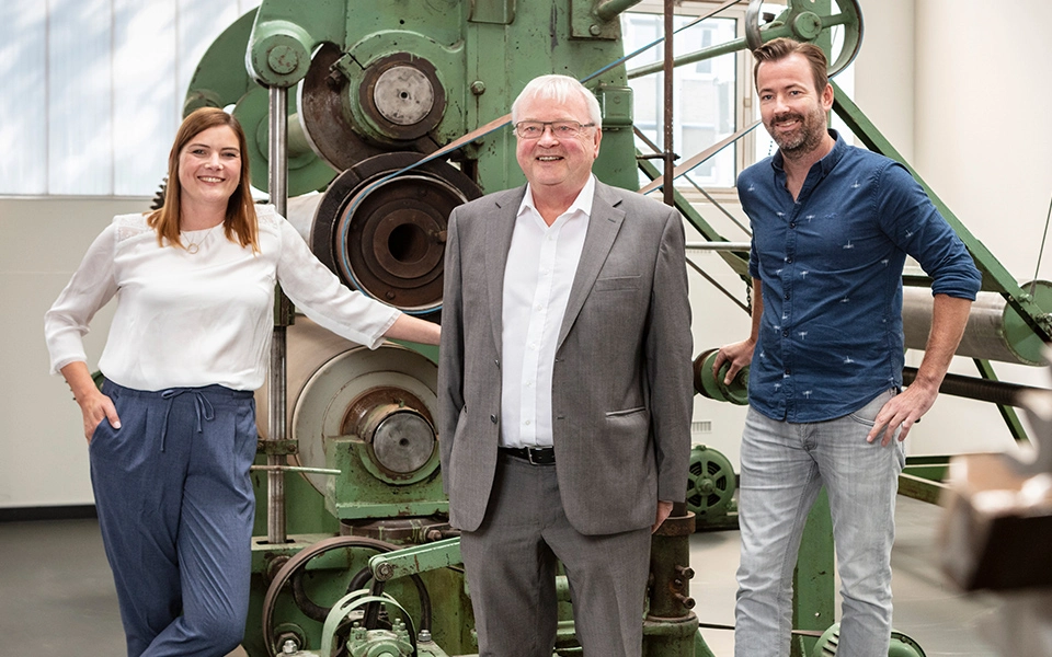 Heinz-Friedrich Kammen mit seinen Kindern Tim Kammen und Nina Sieben, die vor einer historischen Maschine abgebildet sind.