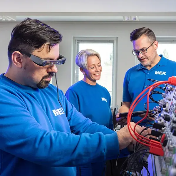 Ausbilder und Teilnehmerin in der Ausbildungswerkstatt für Pneumatik schauen gemeinsam auf ein Tablet, während ein junger Mann im Vordergrund an einer pneumatischen Schaltung arbeitet.