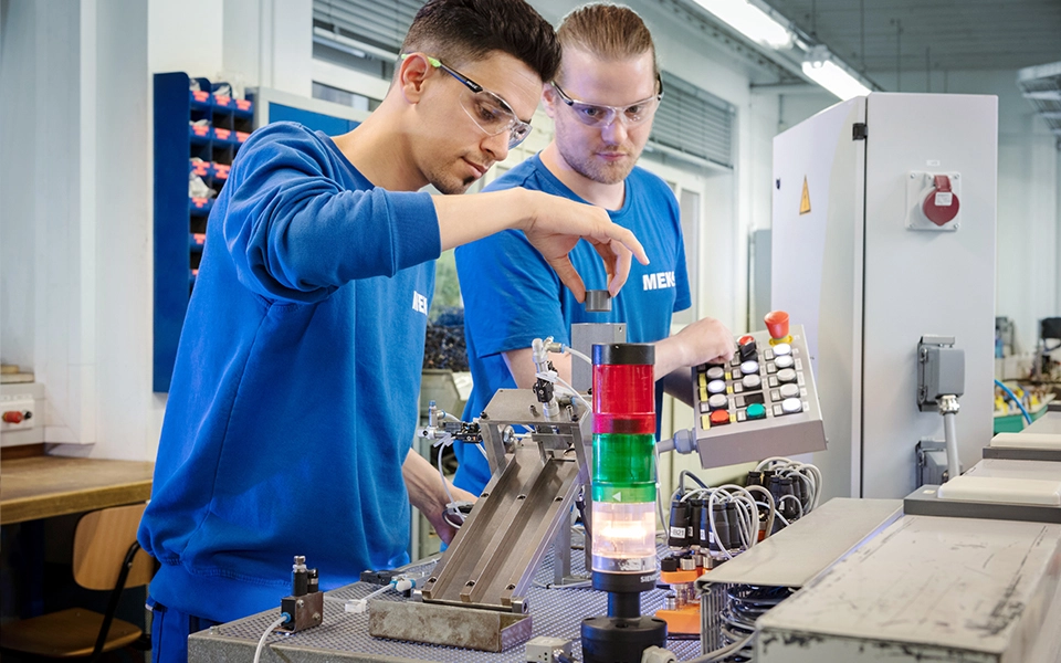 Zwei junge Männer in MEK-Arbeitskleidung arbeiten zusammen an einem Modell in einer Ausbildungswerkstatt.